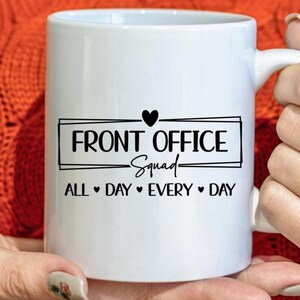Puede incluir: Taza de cer&aacute;mica blanca con el texto "FRONT OFFICE Squad" en fuente negra, con un coraz&oacute;n negro encima del texto. Debajo del texto est&aacute; "ALL DAY EVERY DAY" en fuente negra. La taza tiene un asa blanca.