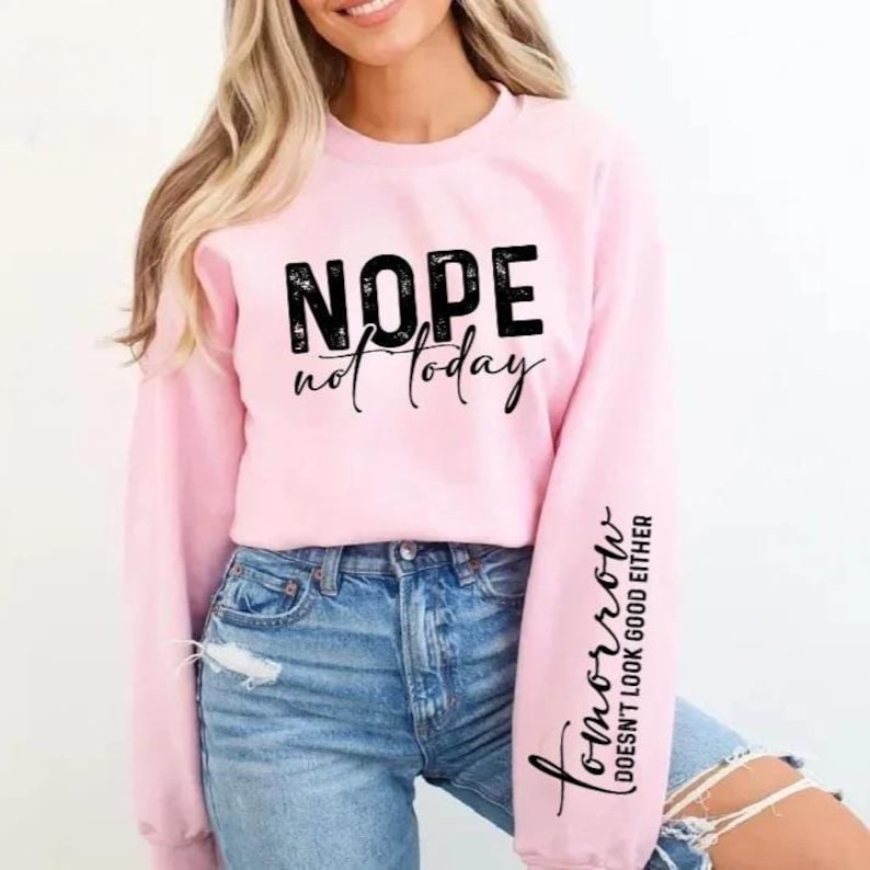 Pode incluir: Camisola rosa claro com as palavras "NOPE not today" impressas em preto na frente. A manga direita tem as palavras "Tomorrow doesn't look good either" impressas em preto. A camisola &eacute; combinada com jeans azuis.