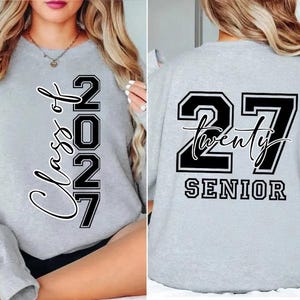 May include: A light gray sweatshirt with the text "Class of 2027" in a vertical script on the front. The back of the sweatshirt features the number "27" with the word "Twenty" and "SENIOR" below it, all in black and white.