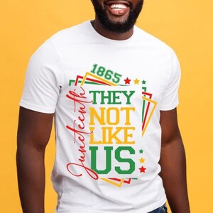 May include: White t-shirt with a colorful graphic design. The design includes the text "1865", "They Not Like Us", and "Juneteenth" in red, green, and yellow. The shirt has a crew neck and short sleeves.