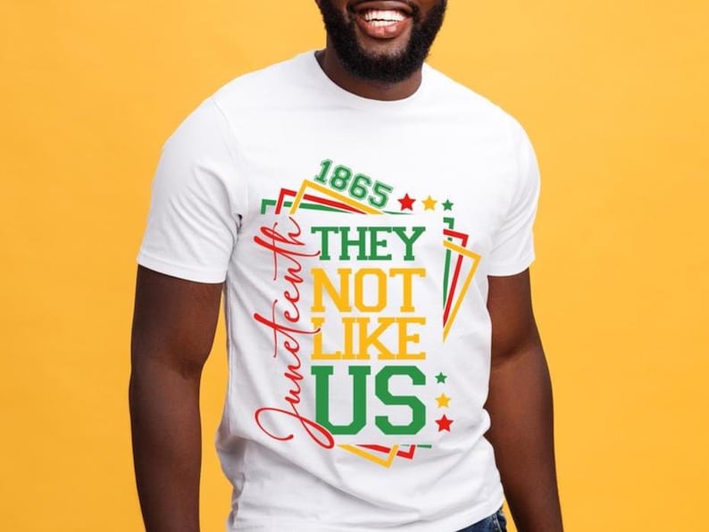 May include: White t-shirt with a colorful graphic design. The design includes the text "1865", "They Not Like Us", and "Juneteenth" in red, green, and yellow. The shirt has a crew neck and short sleeves.