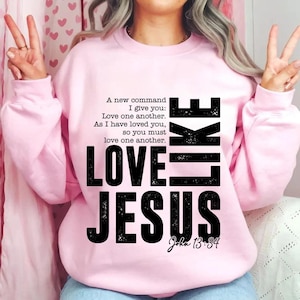 May include: A light pink sweatshirt with the words "LOVE LIKE JESUS" in large black letters. The text includes a quote from John 13:34, emphasizing love and compassion. The sweatshirt is a comfortable, casual style.