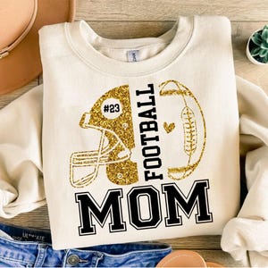 May include: Cream-colored sweatshirt with a gold glitter football helmet and football design. The text "FOOTBALL MOM" is in black block letters. The helmet has the number "23" in a circle.