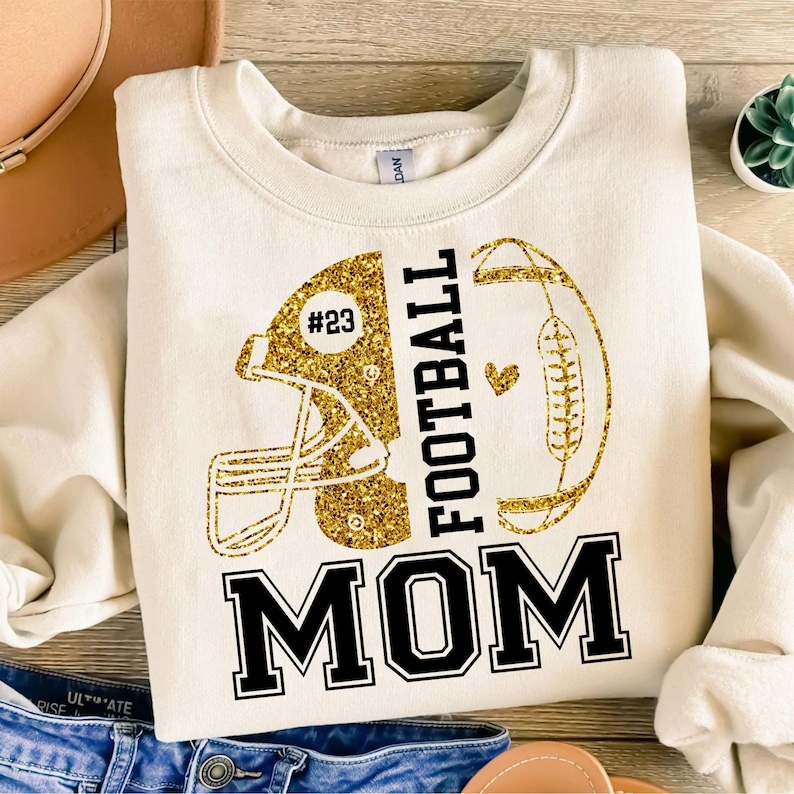 May include: Cream-colored sweatshirt with a gold glitter football helmet and football design. The text "FOOTBALL MOM" is in black block letters. The helmet has the number "23" in a circle.