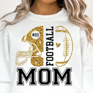 May include: White sweatshirt with a gold glitter football helmet and football design. The word "FOOTBALL" is printed vertically in black, and "MOM" is printed in bold black letters. The helmet has the number "23".