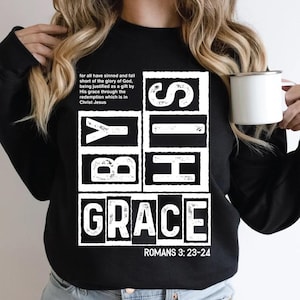 May include: Black sweatshirt with the words "BY HIS GRACE" in a white, bold font. The text is arranged in a vertical block design. Additional text includes a Bible verse reference and a quote about redemption.
