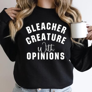 May include: Black sweatshirt with the text "BLEACHER CREATURE With OPINIONS" in white block letters. The word "With" is in a cursive font. A white mug is held in the right hand.
