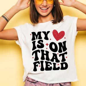 May include: White t-shirt with the text "MY IS ON THAT FIELD" in a bold, black, retro font. A red glitter heart and small red starbursts accent the design. The shirt is a casual, everyday wear item.