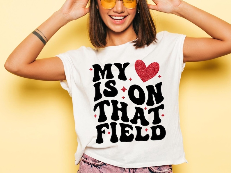 May include: White t-shirt with the text "MY IS ON THAT FIELD" in a bold, black, retro font. A red glitter heart and small red starbursts accent the design. The shirt is a casual, everyday wear item.