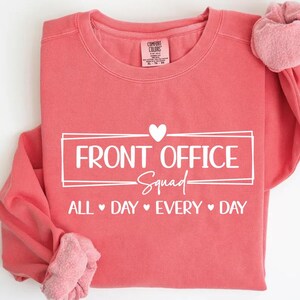 Puede incluir: Sudadera de color coral con texto blanco "FRONT OFFICE Squad ALL DAY EVERY DAY". El dise&ntilde;o incluye un s&iacute;mbolo de coraz&oacute;n. La sudadera tiene cuello redondo y mangas largas. La tela parece suave.