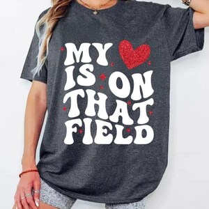 May include: Dark gray t-shirt with the text "MY HEART IS ON THAT FIELD" in white, bold, bubble letters. A red glitter heart is above the word "ON". The shirt is a casual style.