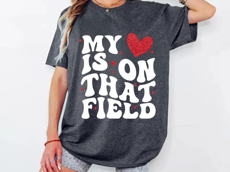 May include: Dark gray t-shirt with the text "MY HEART IS ON THAT FIELD" in white, bold, bubble letters. A red glitter heart is above the word "ON". The shirt is a casual style.