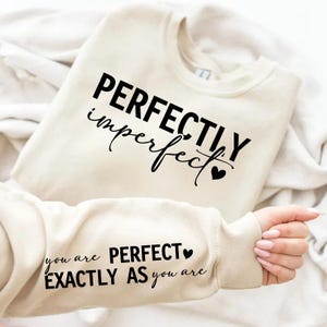 May include: A cream-coloured sweatshirt with the words "PERFECTLY imperfect" printed in black script. The sleeve displays the text "you are PERFECT EXACTLY AS you are" in black. A black heart is next to the word perfect.
