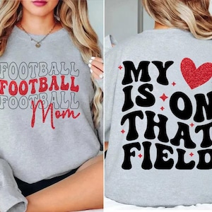 May include: Light gray sweatshirt with the words "FOOTBALL Mom" in red glitter and black lettering on the front. The back of the sweatshirt says "MY IS ON THAT FIELD" with a red glitter heart.