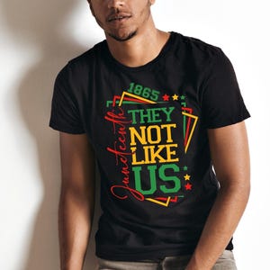 May include: Black t-shirt with a colorful graphic design. The design includes the text "1865" and "They Not Like Us" in green, yellow, and red. The word "Juneteenth" is written in a cursive font.
