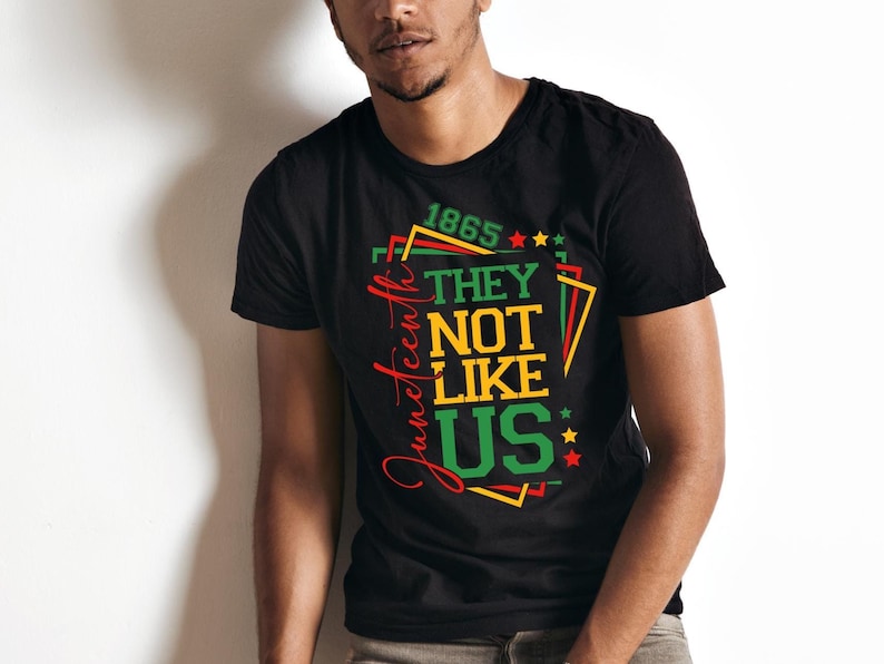 May include: Black t-shirt with a colorful graphic design. The design includes the text "1865" and "They Not Like Us" in green, yellow, and red. The word "Juneteenth" is written in a cursive font.