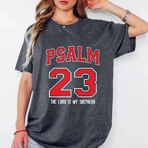 May include: Dark gray t-shirt with the text "PSALM 23" in red letters with a white outline, and the number "23" in red with a white outline. Below the number is the text "THE LORD IS MY SHEPHERD".