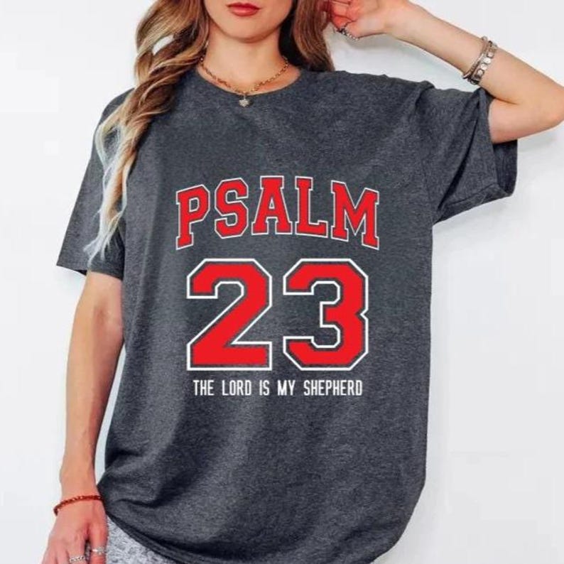 May include: Dark gray t-shirt with the text "PSALM 23" in red letters with a white outline, and the number "23" in red with a white outline. Below the number is the text "THE LORD IS MY SHEPHERD".