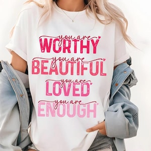 May include: White t-shirt with the phrases "You are WORTHY, BEAUTIFUL, LOVED, ENOUGH" in shades of pink. The text is arranged in a stacked format with decorative hearts. The shirt has short sleeves and a crew neck.