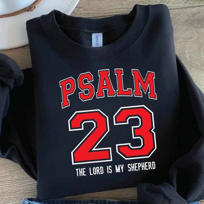 May include: Black sweatshirt with the text "PSALM 23" in red block letters with a white outline. Below the number "23" is the phrase "THE LORD IS MY SHEPHERD" in white. The sweatshirt is folded.