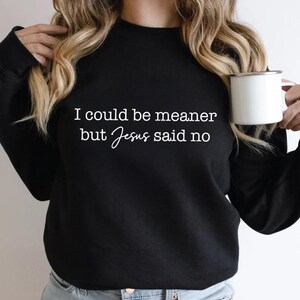 May include: Black sweatshirt with the text "I could be meaner but Jesus said no" in white. The sweatshirt is a crew neck style. A white mug is held in the right hand.