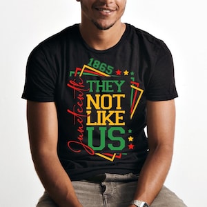 May include: Black t-shirt with a colorful graphic design. The design includes the text "1865", "Juneteenth", and "THEY NOT LIKE US" in red, green, yellow, and black. The shirt is a crew neck style.