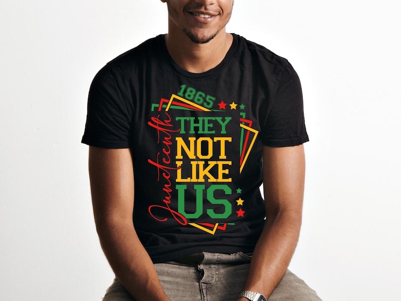 May include: Black t-shirt with a colorful graphic design. The design includes the text "1865", "Juneteenth", and "THEY NOT LIKE US" in red, green, yellow, and black. The shirt is a crew neck style.