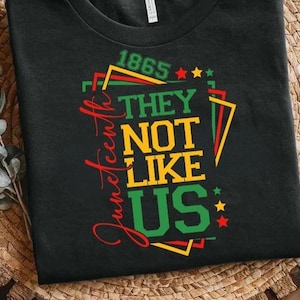 May include: Black t-shirt with the text "Juneteenth" in red script. The shirt features the phrase "THEY NOT LIKE US" in yellow and green, with the year "1865" above. Stars and geometric shapes in red, green, and yellow complete the design.