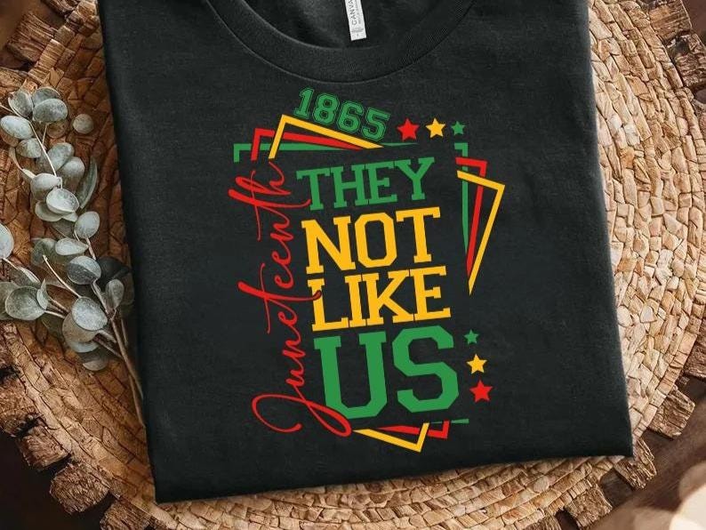 May include: Black t-shirt with the text "Juneteenth" in red script. The shirt features the phrase "THEY NOT LIKE US" in yellow and green, with the year "1865" above. Stars and geometric shapes in red, green, and yellow complete the design.