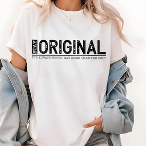 May include: White t-shirt with the words "STAY ORIGINAL" in black block letters. Below the main text is the phrase "IT'S ALWAYS WORTH WAY MORE THAN THE COPY". The shirt is a casual style.