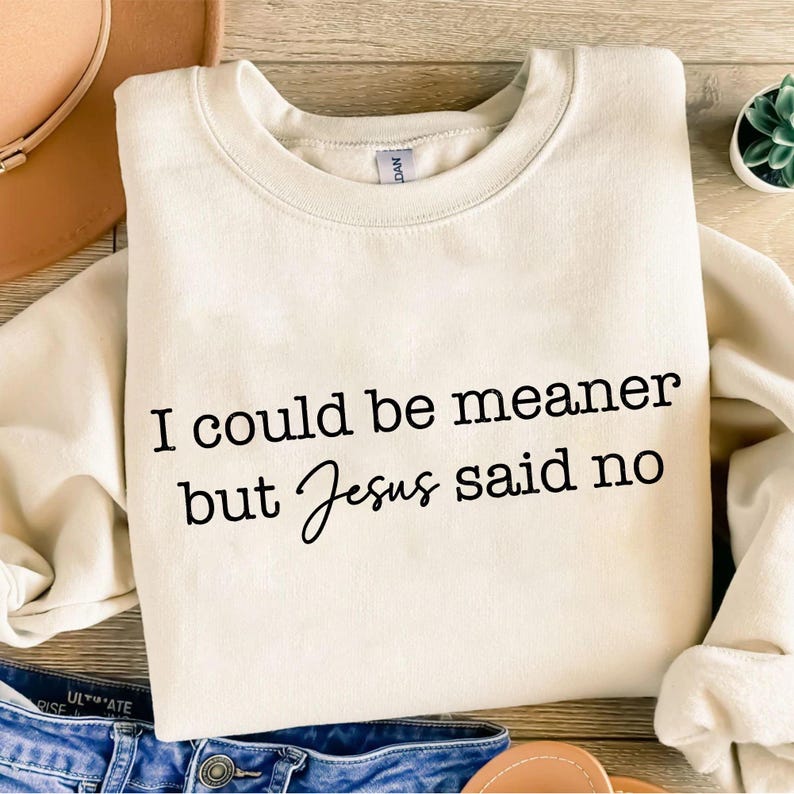May include: Cream-colored sweatshirt with the text "I could be meaner but Jesus said no" in black. The sweatshirt is folded, with a hint of denim jeans and a hat in the background.