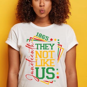 May include: White t-shirt with a colorful graphic design. The design includes the text "Juneteenth" in red script, and the words "THEY NOT LIKE US" in green and yellow. The number "1865" is also included.