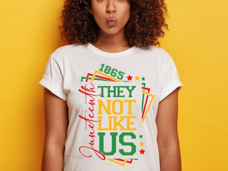 May include: White t-shirt with a colorful graphic design. The design includes the text "Juneteenth" in red script, and the words "THEY NOT LIKE US" in green and yellow. The number "1865" is also included.