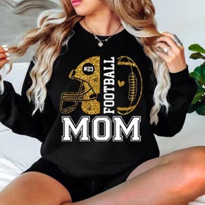 May include: Black sweatshirt with a gold glitter football helmet and football design. The text "FOOTBALL MOM" is printed in white block letters. The helmet has the number "23" on it.