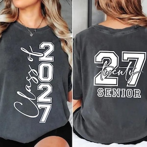 May include: Charcoal gray t-shirt with white text. The front displays "Class of 2027" vertically. The back features "27 Twenty SENIOR" in a bold, collegiate style. A casual, comfortable tee for graduation.