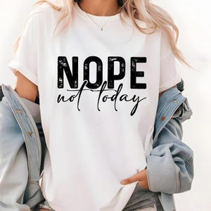Pode incluir: Camiseta branca com a frase "NOPE not today" em preto. A palavra "NOPE" est&aacute; em negrito, com um estilo desgastado, e "not today" em it&aacute;lico. A camiseta tem mangas curtas.