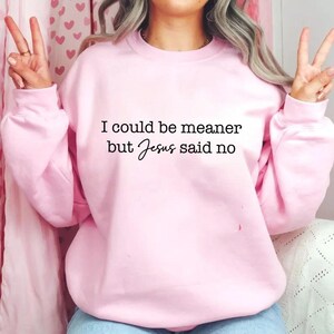 May include: A light pink sweatshirt with the text "I could be meaner but Jesus said no" in black. The sweatshirt has long sleeves and a crew neck.