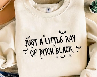 Puede incluir: Sudadera color crema con la frase "Just a Little Ray of Pitch Black" en negro, junto con gráficos de murciélagos, arañas y estrellas. La sudadera de cuello redondo es de estilo casual.