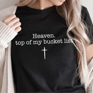 May include: Black t-shirt with the text "Heaven. top of my bucket list" in white, along with a white cross graphic. The shirt is being worn with a cream-colored cardigan.