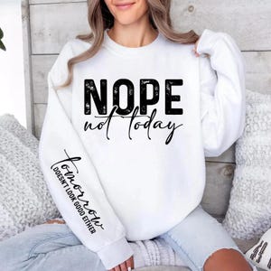 Pode incluir: Camisola branca com as palavras "NOPE not today" em preto, com fonte desgastada. A manga apresenta o texto "Tomorrow doesn't look good either" em uma fonte cursiva. A camisola &eacute; um estilo casual e confort&aacute;vel.