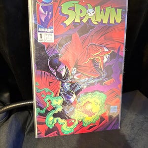 Pode incluir: Uma capa de banda desenhada Spawn com um personagem vermelho e preto com uma caveira no peito. A banda desenhada é o número 1 e intitula-se "Spawn".