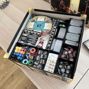 May include: A board game organizer with various compartments filled with game components. Includes cards, dice, tokens, and game boards. The organizer is black with a yellow trim and is designed to hold all the game pieces.