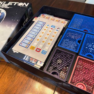 May include: Board game box titled "Shackleton Base" with an astronaut illustration. The box is open, revealing game cards, blue and purple card holders, and a red card holder. The game components are neatly arranged.