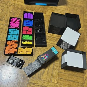 Puede incluir: Un juego de mesa con formas geom&eacute;tricas coloridas, bandejas negras y cartas. El juego incluye varias piezas de rompecabezas de colores, piezas de juego negras y una caja con un acento azul. El juego est&aacute; sobre una superficie de madera.