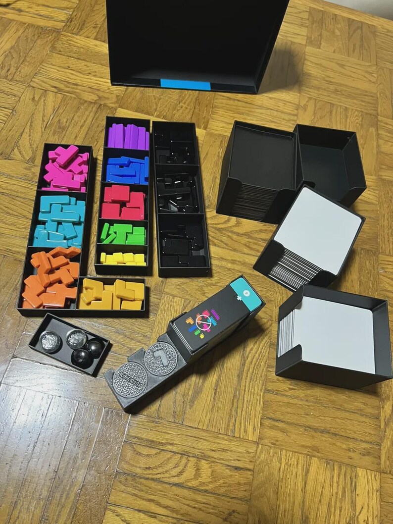 Puede incluir: Un juego de mesa con formas geom&eacute;tricas coloridas, bandejas negras y cartas. El juego incluye varias piezas de rompecabezas de colores, piezas de juego negras y una caja con un acento azul. El juego est&aacute; sobre una superficie de madera.