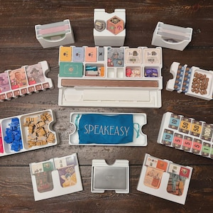 May include: A board game organizer with various white plastic trays and containers filled with game components. Includes cards, tokens, dice, and a blue bag with the word "SPEAKEASY". The game pieces are arranged on a dark wooden surface.