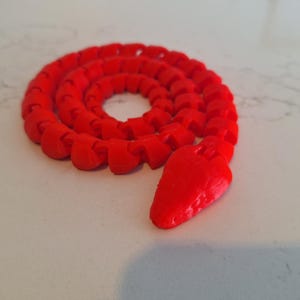 May include: A bright red, segmented snake toy made of plastic. The snake is curled up in a circle with its head pointing upwards.