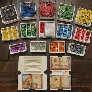 以下が含まれることがあります： 白いトレイに配置された、布の切れ端、カラフルなトークン、ゲームカードなど、ボードゲームのコンポーネントのコレクション。 コンポーネントは、青、緑、赤、黄色などのさまざまな色で、濃い木製の表面に配置されています。