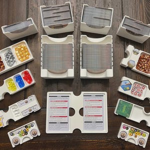 May include: A collection of white plastic game organizers and card holders, filled with game pieces and cards. The organizers are arranged on a dark wood surface, showcasing various game components in different colors and shapes.