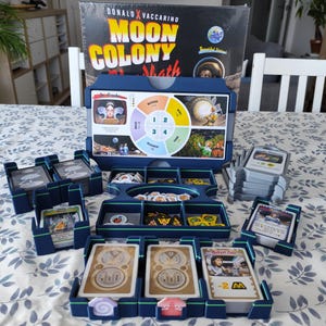 May include: A board game titled "Moon Colony" by Donald X. Vaccarino. The game box and components are arranged on a table. The game includes cards, tokens, and a game board with a circular diagram. The color scheme is primarily blue, white, and gold.